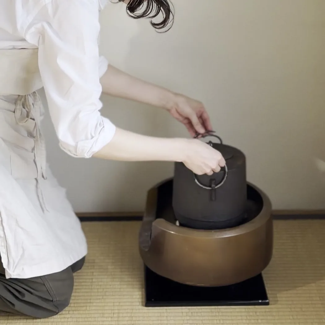 割烹着の人が、炉の上の釜に手を伸ばしている